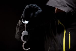A criminal in a hood has handcuffs on one wrist. 